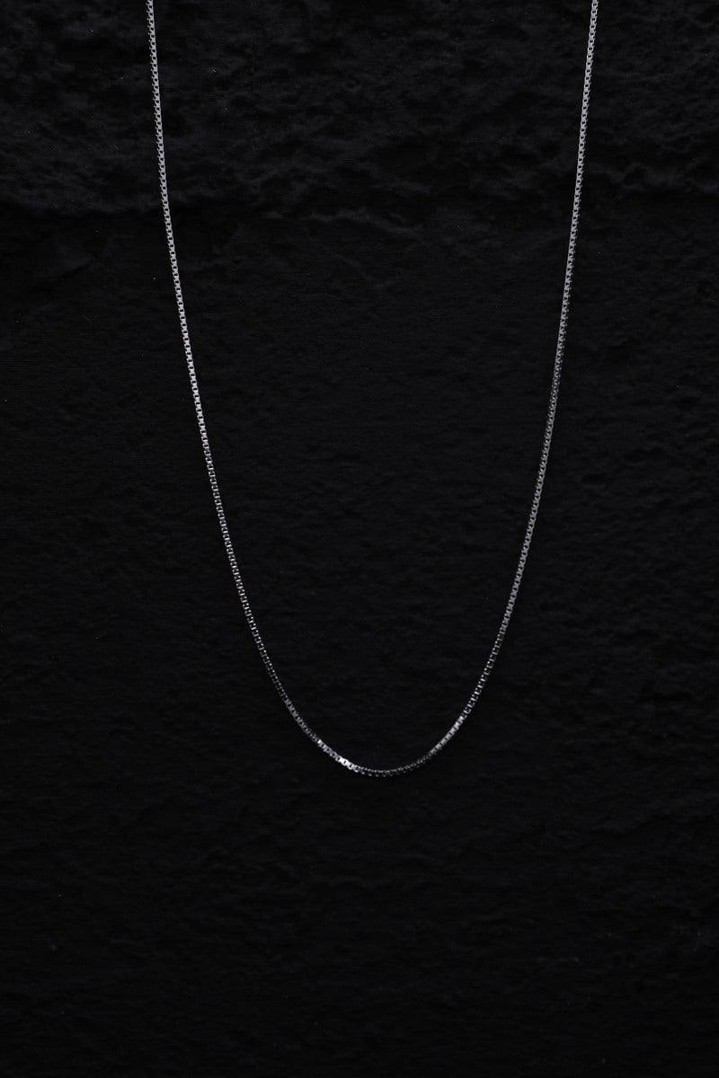 Close-up of a delicate silver box chain necklace shimmering against a black background. Classic, versatile design.