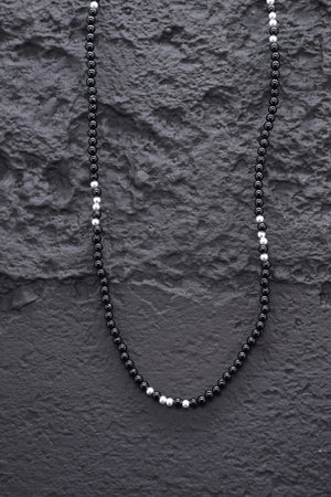 Close-up of a minimalist black onyx beaded necklace with silver accents on a textured black background.