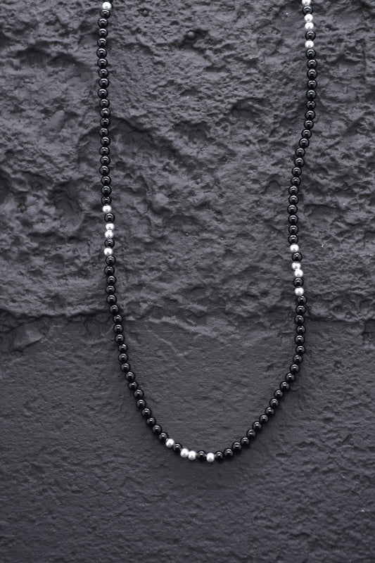 Close-up of a minimalist black onyx beaded necklace with silver accents on a textured black background.