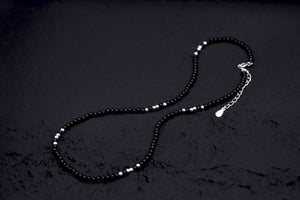 Elegant minimalist necklace with small black and silver Tibetan dzi beads on a textured black background.