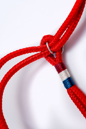 Close-up of a red braided rope bracelet with burgundy, white, and teal accents. The metal ring closure and textured weave add a unique touch to this Buddhist prayer beads-inspired accessory.