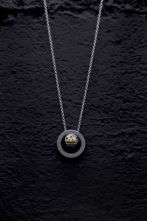 Silver chain necklace with dark circular pendant featuring a gold triangle, eye, and diamond. Minimalist, gothic dzi bead pendant on textured black background.
