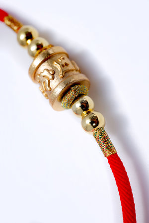 Close-up: Red cord bracelet with gold prayer wheel pendant. Embossed details, gold beads, and colorful thread wraps. Buddhist prayer wheel for meditation.