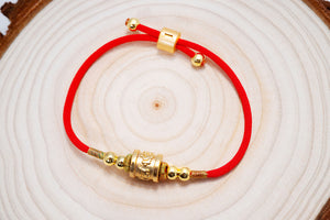 Red string bracelet with gold beads and etched charm on wood. Tibetan dzi beads add vibrant color and detail.