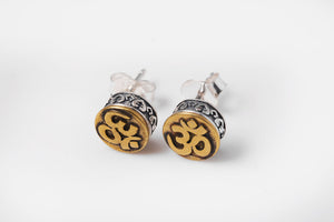 Close-up of two Om symbol stud earrings. Gold Om on a circular gold background, framed by etched silver. Simple, detailed Buddhist prayer jewelry.