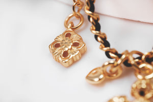 Close-up of gold floral charm bracelet with black cord, showcasing ornate pendants. Tibetan dzi beads add a unique touch.