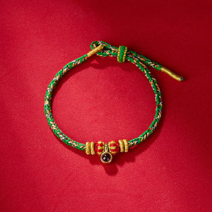 Braided bracelet with green, red, gold threads, sliding knot, and dark gemstone on vibrant red. Handmade jewelry.