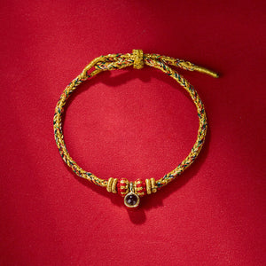 Braided bracelet featuring gold thread with red, green, and blue strands, red beads, and a dark stone charm.
