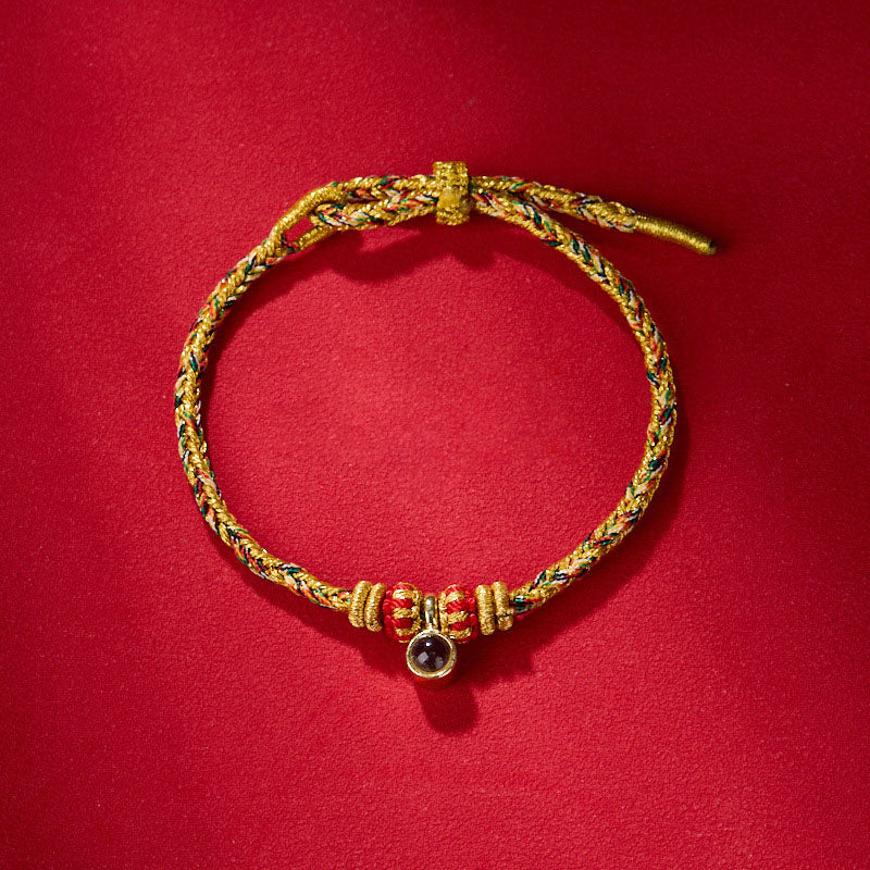 Braided bracelet featuring gold thread with red, green, and blue strands, red beads, and a dark stone charm.