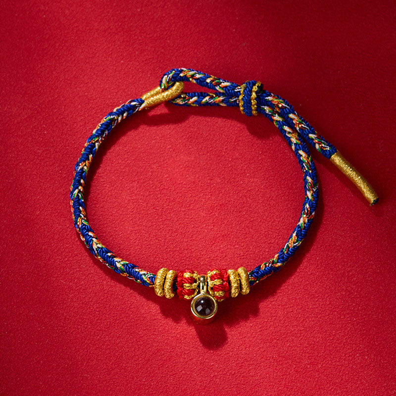 Braided bracelet with blue, red, yellow, and white cords, featuring a stone pendant with gold accents. Handcrafted jewelry on a red fabric.
