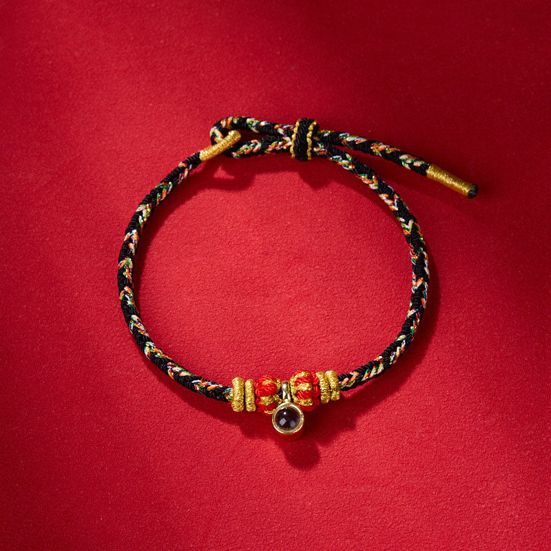 Braided bracelet with red, green, and gold threads, textured gold and red beads, and gemstone accent.