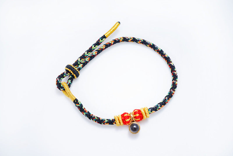 Braided cord bracelet with red, green, and yellow threads, gold accents, knotted beads, and a dark gemstone. Bohemian style jewelry.