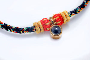 Braided bracelet close-up: black, red, yellow, green threads with gold accents and a dark blue stone pendant.