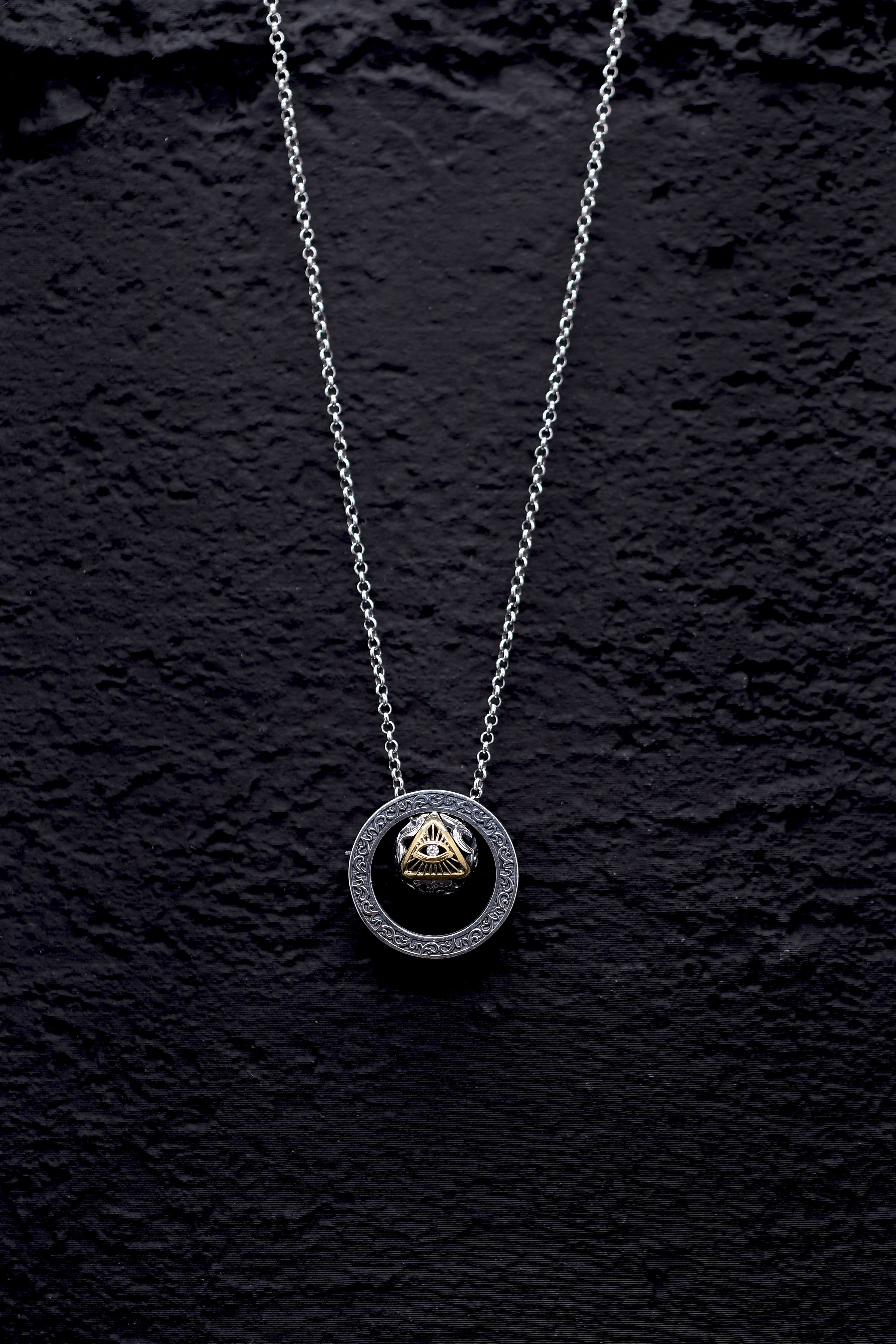 Silver chain necklace with dark circular pendant featuring a gold triangle, eye, and diamond. Minimalist, gothic dzi bead pendant on textured black background.