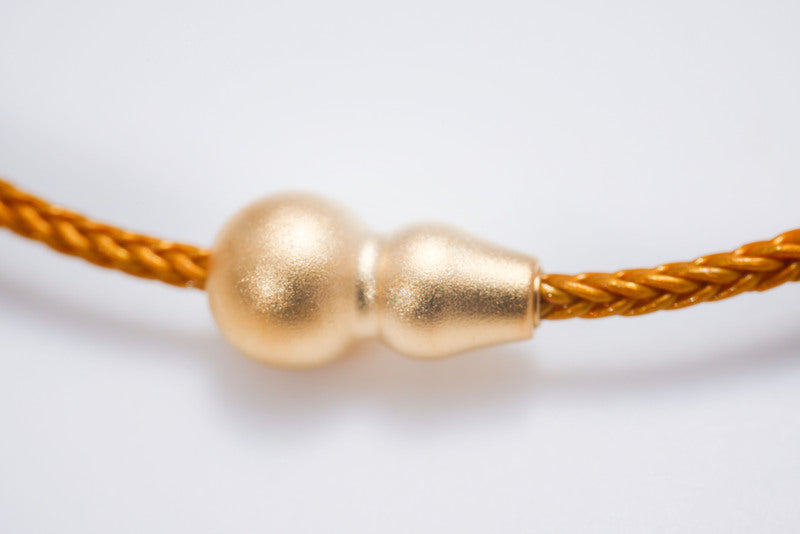Close-up of a matte gold gourd-shaped Tibetan dzi bead on a braided gold cord. Minimalist style, perfect for Tibetan dzi stone beads lovers.