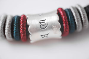 Close-up of braided bracelet with silver "WB" Tibetan dzi bead. Gray, teal, & maroon interwoven strands.