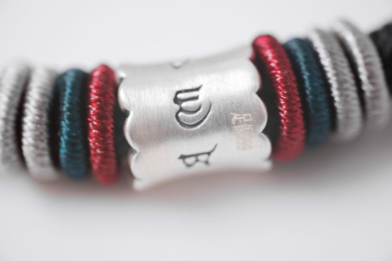 Close-up of braided bracelet with silver "WB" Tibetan dzi bead. Gray, teal, & maroon interwoven strands.