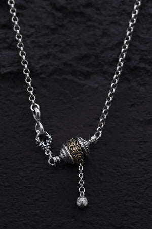 Close-up of silver & gold Tibetan prayer wheel pendant necklace on black background. Etched details, dangling bead, decorative clasps. Buddhist jewelry.