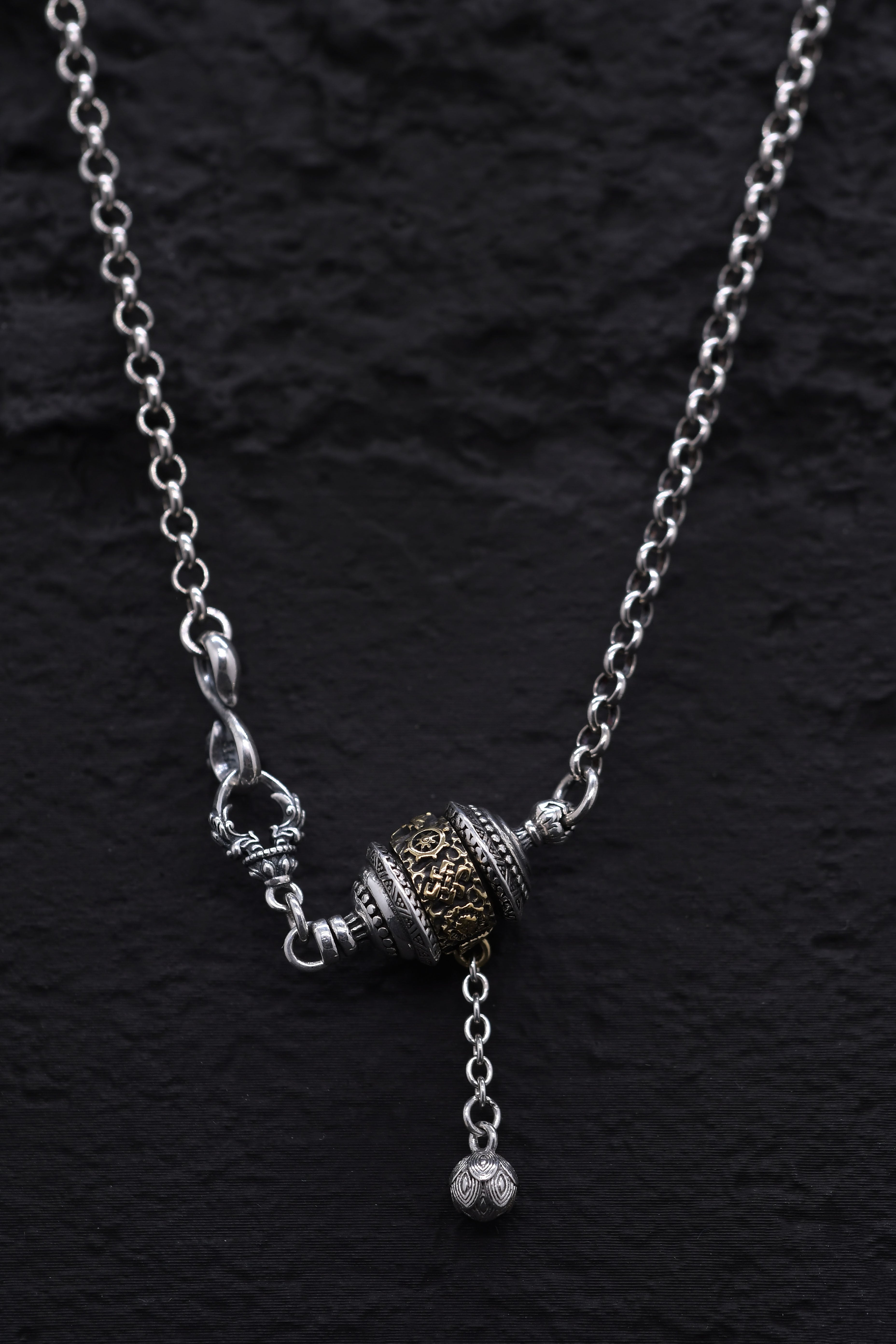 Close-up of silver & gold Tibetan prayer wheel pendant necklace on black background. Etched details, dangling bead, decorative clasps. Buddhist jewelry.