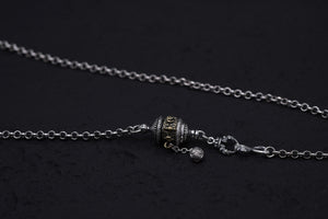 Close-up of a silver and gold Tibetan prayer wheel pendant necklace on a textured black background. Intricate engravings and a dangling bead add to this unique Buddhist prayer wheel's detail.