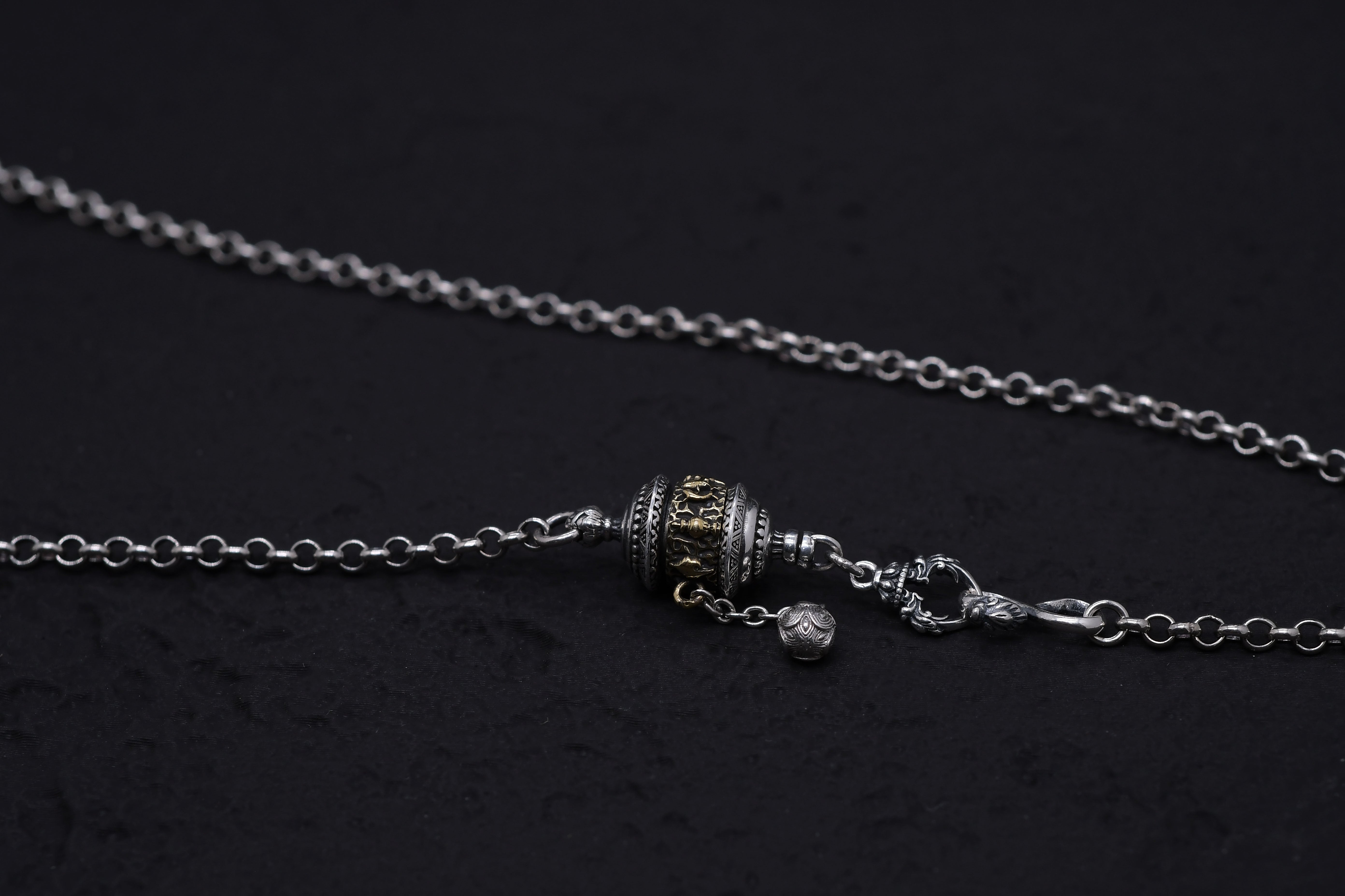 Close-up of a silver and gold Tibetan prayer wheel pendant necklace on a textured black background. Intricate engravings and a dangling bead add to this unique Buddhist prayer wheel's detail.