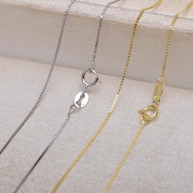 Close-up of delicate silver and gold box chain necklaces. Simple background emphasizes the clean, product-focused style. Amulet necklace options for layering.