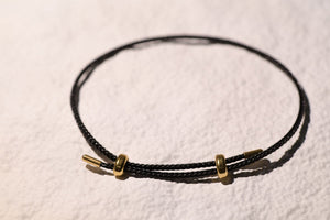 Black braided bracelet with gold rings and cylindrical ends on textured surface. Add personalized charms to this woven stainless steel string bracelet.