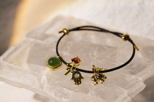 Black cord bracelet with green gemstone, key, Hamsa, elephant, and reddish-orange charms.