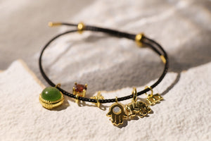 Black cord charm bracelet with green jade, red garnet, star, hamsa hand, elephant, and "M" charms.