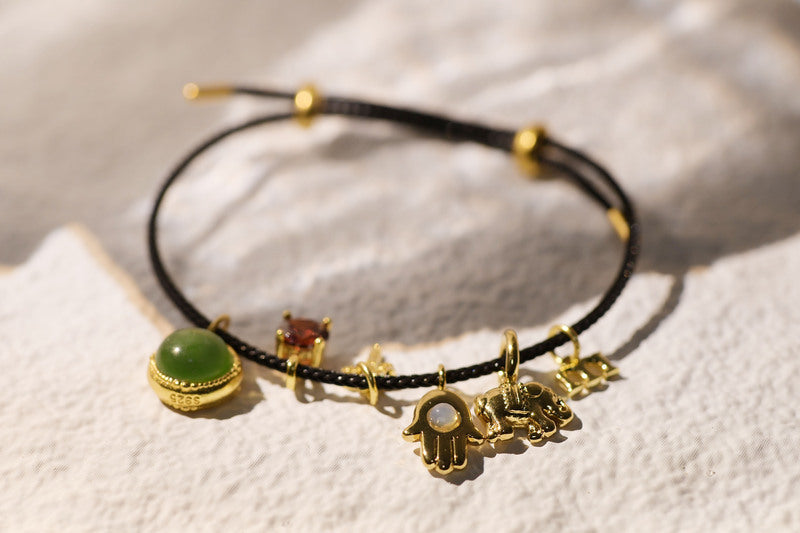 Black cord charm bracelet with green jade, red garnet, star, hamsa hand, elephant, and "M" charms.