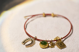 Red string bracelet with horseshoe, jade, Om, and star charms on a textured surface.