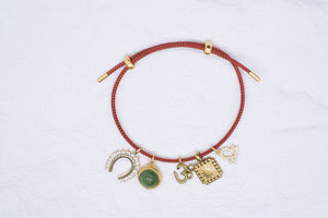Red braided charm bracelet featuring horseshoe, jade pendant, Om symbol, sunburst, and heart charms. Gold accents. 