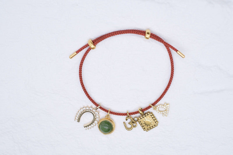 Red braided charm bracelet featuring horseshoe, jade pendant, Om symbol, sunburst, and heart charms. Gold accents. 